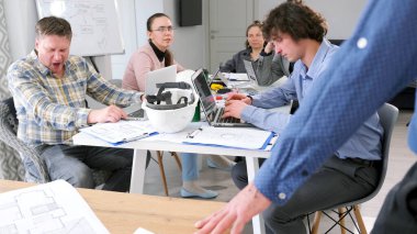 Küçük bir inşaat şirketinde brifing. Bir grup ofis çalışanı kağıt ve dizüstü bilgisayarlarla dolu bir masada oturuyor. Sahne odaklanmış ve ciddi. Şirket personeli proje üzerinde çalışıyor.