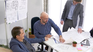 Üç adam inşaat planlarını tartışıyor. İş adamı proje bütçesini mimar ve mühendis ile koordine ediyor. Masada çizimler, belgeler ve diğer kağıtlar var..