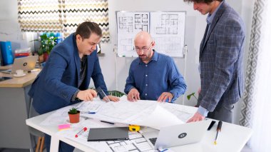 Üçgen cetvelli bir adam çizimlere işaret eder ve mimardan tesisin inşaat zamanını sorar. Toplantı odasında masada üç tane iş adamı var. Proje bütçesinin tartışılması