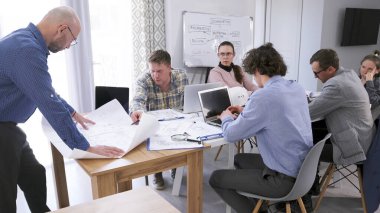 Mimar çizimde kalemi ile işaret eder. Bir grup ofis çalışanı, arkasında diyagramı olan beyaz bir tahtayla bir masada oturuyorlar. Birlikte bir proje üzerinde çalışıyorlar..