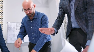 Gözlüklü ve elinde kalem olan mavi gömlekli bir adam çizimde değişiklikler yapar. Müşteri yerleşim planında düzenlemeler yapar. Adamlar Mimarlık Bürosu 'nun konferans salonunda işbirliği yapıyorlar..