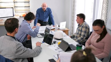 Sabah planlama seansı. Bir grup iş adamı konferans odasında bir masada oturuyor. Merkezdeki adam bir sunum yapıyor. Sahne profesyonel ve odaklı.