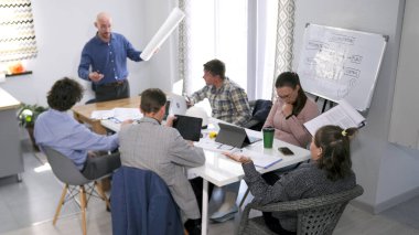 Patron, çalışanlarına yapılan iş hakkında rapor sorar. Bir grup insan konferans odasında bir masada oturuyor. Bir adam çizimleriyle grubun önünde duruyor ve bir sunum yapıyor..