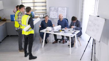 Bir inşaat işçisi, bir mühendis ve bir tasarımcı toplantı odasında aynı masada oturmuş yeşil yelekli iki işçiyle sohbet ediyorlar. Bir grup adam bir konferans odasında toplanıp proje planlarını tartıştılar..