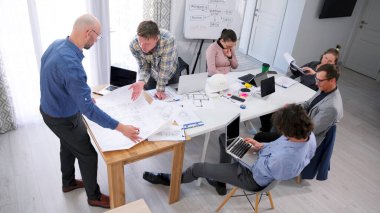 Bir grup insan konferans odasında dizüstü bilgisayarlar ve belgelerle bir masada oturuyor. Proje çizimlerinin tartışılması. Bir şirket, bir inşaat şirketi için reklam geliştiriyor..