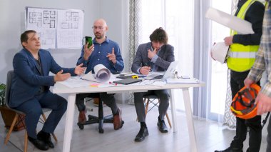 İnşaatçıların sorularını üç işadamı cevaplıyor. Bir grup adam konferans odasında bir masada oturmuş proje tahminleri, inşaat aşamaları ve çalışmanın diğer ayrıntılarını tartışıyorlar..