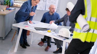 İnşaat şirketinde brifing. Ustabaşı, mimar ve mühendislerle toplantı. Bir grup insan çizimler ve kağıtlarla bir masada oturuyor. Yeşil yelekli bir işçi bina planıyla geldi..