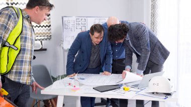 Ustabaşı proje çizimlerini yönetimle koordine edecek. Bir grup insan, planları ve dizüstü bilgisayarı olan bir masanın etrafında toplandı. Proje için planları ve fikirleri tartışıyorlar..