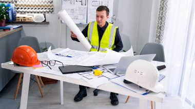 Yeşil sinyal yeleği giymiş genç bir ustabaşı, önünde çizimler, planlar, dizüstü bilgisayar ve miğfer olan bir masada oturuyor. Bir inşaat mühendisi ofisinde çalışıyor.