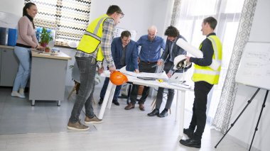 Ustabaşı, müteahhit ve bir grup müşteri proje çizimlerini tartışıyor. İnşaat şirketinde bir toplantı. Bir grup insan rahat bir ofiste masanın etrafında dikiliyorlar..