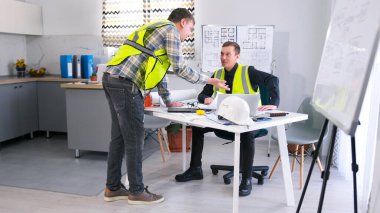 Mimari ofis. Yeşil yelekli iki erkek işçi bir proje üzerinde çalışmaktan bahsediyor. Ustabaşı ve müteahhit projenin tahmini ve detaylarını tartışıyor..