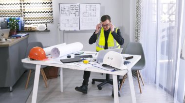 Koruyucu yeşil yelek ve gözlüklü genç bir adam bir dizüstü bilgisayar, çizimler ve bir yığın kağıtla bir masada oturuyor. Masanın yanında inşaat kaskları, mezura, telefon vs. var..