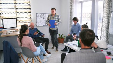 Elinde mavi klasör olan bir adam bir iş merkezi inşaatı sunumunu yönetiyor. Bir grup insan, projenin tahminlerini ve detaylarını tartışmak üzere bir konferans odasında toplandı..