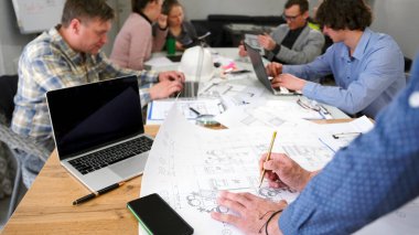 Mimarın ellerine yakın plan çizimlerinde değişiklikler yapıyor. Bir grup insan akıllı bir ev inşaatı projesinde birlikte çalışıyor. Laptoplar, kağıtlar ve kalemlerle dolu bir masada oturuyorlar..