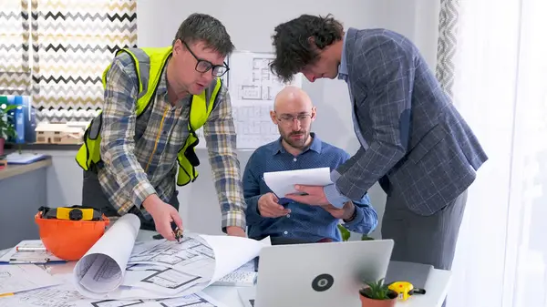 Üç odaklı adam, modern bir iş merkezi projesinin çizimleri, planları ve belgeleriyle çalışıyor. Mimarlar proje tahminlerini tartışıyor ve her şeyin yolunda olduğundan emin oluyorlar