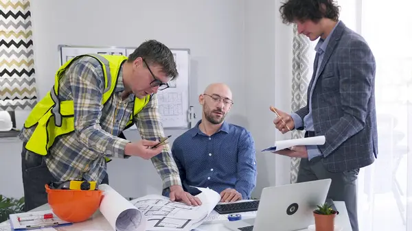 Mimarlar, inşaatçılar ve mühendislerden oluşan profesyonel bir ekip modern bir binanın çizimleri ve planları üzerinde çalışıyor. Üç adam inşaatın ayrıntılarını ve aşamalarını tartışıyor..