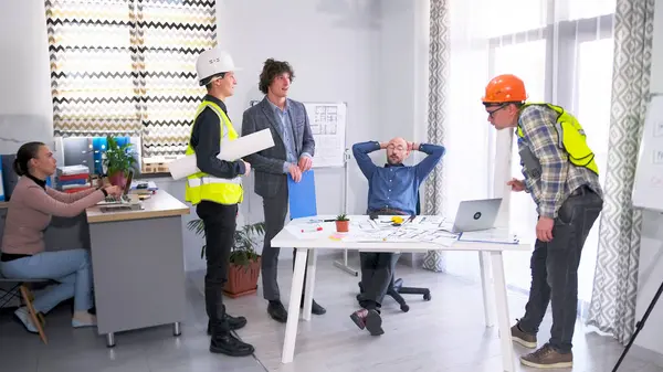 Bir grup inşaat işçisi, bir mühendis ve bir mimar çizimlerle dolu bir masanın etrafında toplandılar. İşçiler planları tartışırken sahne işbirliği ve takım çalışmasını temsil ediyor.