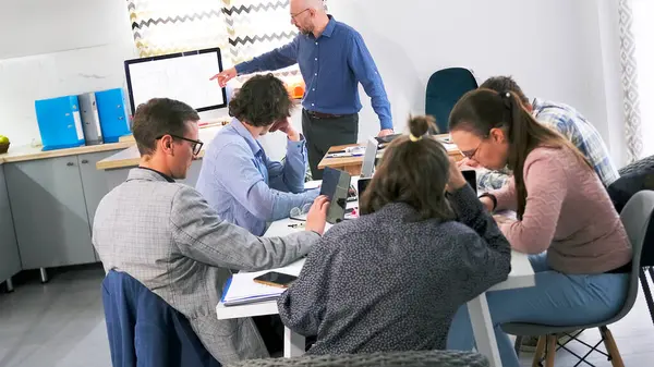 Bir grup insan toplantı odasında bir masada oturuyorlar. Bir adam önlerinde duruyor ve bir sunum yapıyor. Sahne profesyonel ve odaklanmış. Brifing ve takım çalışması
