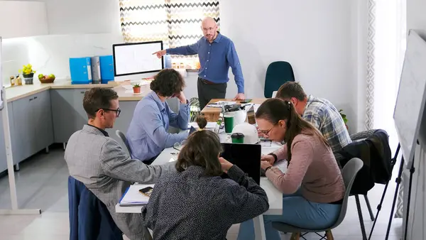 Bir grup insan konferans odasında bir masada oturuyor ve bir sunum izliyor. Adam ekranı işaret ediyor. Toplantı ya da brifing. Sahne odaklı ve ciddi, grup bir proje üzerinde çalışıyor.