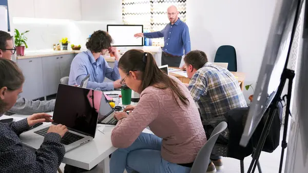 Bir grup öğrenci dizüstü bilgisayarları açık bir masada oturuyorlar. Bir adam grubun önünde duruyor ve ekrandaki bir şeyi işaret ediyor. İnşaatla ilgili bir konferansta sunum. Sahne ortada.