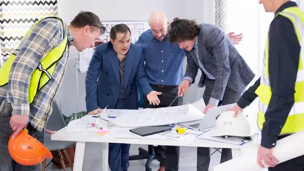 Bir grup adam elinde planlarla bir masanın etrafında toplandı. Akıllı ev projesinin tartışması. Yeşil yelekli işçiler ve iş adamları projenin tahminleri ve ayrıntıları hakkında konuşuyor