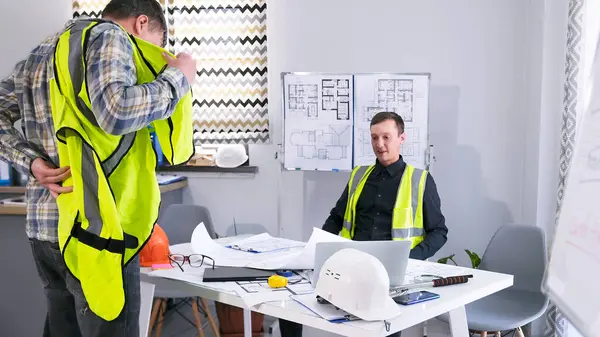 Bir erkek işçi ustabaşının ofisinde yeşil yelek giyiyor. İki adam bir inşaat alanı için çalışma planını tartışıyorlar. Müteahhit çizimler, planlar ve dizüstü bilgisayarla bir masada oturuyor.