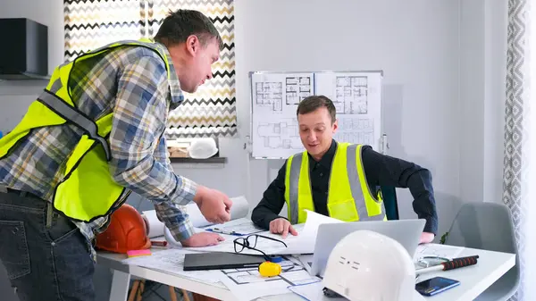 Yeşil yelekli iki işçi inşaat planlarını tartışıyor. Çocuklar çizimlere bakar ve projenin detaylarını tartışırlar. Takım çalışması. Bilgilendirme