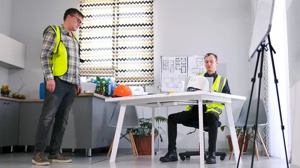 Gözlüklü ve yeşil yelekli bir işçi amirin önünde duruyor ve talimatları bekliyor. İki adam modern bir inşaat projesi üzerinde çalışıyor. Bir mimarlık bürosunun günlük hayatı