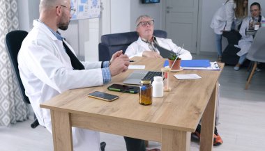 Bir grup doktor personel odasında dinleniyor. Beyaz laboratuvar önlüklü bir adam elinde dizüstü bilgisayar ve bir şişe hapla masada oturuyor. Karşısında başka bir adam oturuyor. Arkada iki stajyer var.