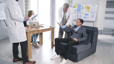 Takım elbiseli bir adam masanın önündeki koltukta oturuyor ve arkasında bir doktor ve bir hemşire var. Yıllık sağlık kontrolü. Tıp ve sağlık hizmetleri kavramı.