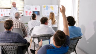 Genç stajyer hocaya bir soru sormak için elini kaldırdı. Bir grup doktor bir odada sandalyede oturuyor ve bir adam önlerinde duruyor ve yeni bir bilimsel çalışma sunuyor..