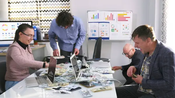 Dört çalışan büyük bir yatırımcıdan para sayıyor. Bir grup insan dizüstü bilgisayarlarla bir masada oturuyor ve her yere para saçıyorlar..