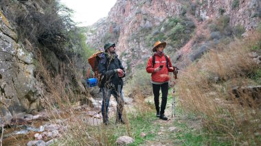Sırt çantalı ve yürüyüş direkli iki yürüyüşçü yeni bir dağ yolunu keşfediyor. İki adam nehir boyunca toprak bir patika boyunca yürüyorlar. Seyahat.