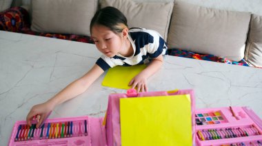Asyalı bir kız güzel bir şey çizmek için renkli kalemlere uzanıyor. Bir çocuk elinde pembe renkli boya kalemleri, boyalar ve bir kağıt parçasıyla masada oturuyor..