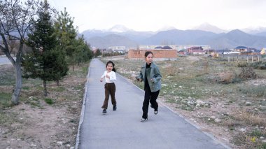 İki kız kardeş kaldırım boyunca koşuyor. Asyalı çocuklar kırsal alanda oynarlar. Çocukların spor ve fiziksel aktiviteleri. Dağlar arka sokakta görünür..