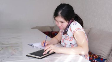 Uzaktan kumanda. Meşgul bir kadın elinde bir defter ve kalemle masada oturuyor. Asyalı bir bayan cep telefonuyla konuşuyor. Ev işleri işte. İş yerinde telefon kullanmak.