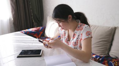 Güzel bir Asyalı kadın bir masada oturuyor cep telefonu, tablet, not defteri ve kalemle. Öğrenci ya da iş kadını telefonuna bakar ve defterine bir şeyler yazar..