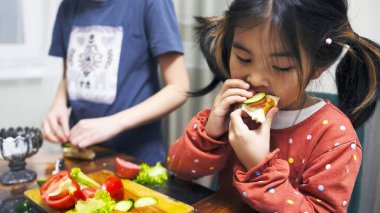 Asyalı tatlı kız bir dilim salatalık ve domatesle sandviç yiyor. Kız kardeşi arkasında duruyor. Çocuklar ev yapımı sandviçleri ev mutfağında yerler.