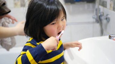 Dişlerini diş fırçasıyla fırçalayan komik bir bebeğin yakın çekimi. Genç bir Asyalı kız banyoda ağız hijyeniyle ilgileniyor. Annesi onun arkasında duruyor..