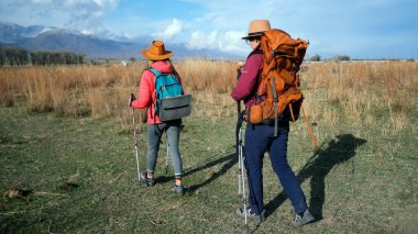 İki gezgin otlak bir arazide yürüyor, her biri sırt çantaları ve yürüyüş direkleriyle. Önümüzde dağlar var. Sahne sakin ve huzurlu.