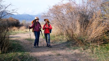 İki turist toprak bir yolda duruyor ve birbirleriyle sohbet ediyorlar. Sırt çantaları ve yürüyüş direkleri olan iki kadın dağlık yabanda uzun bir yürüyüşten sonra dinleniyorlar..