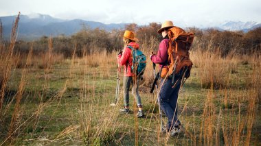 Dağ eteklerinde iki turist yürüyüş yapıyor. İki lezbiyen otlak bir alanda geziyor, her biri şapka takıyor ve sırt çantası taşıyor. Sahne sakin ve huzurlu.
