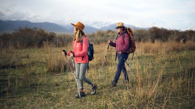 İki yorgun kadın zorlu bir yürüyüş gününden sonra çimenli bir arazide yürüyorlar. Dağ eteklerinde seyahat ediyorum. Sırt çantalı ve sırıklı turistler doğanın tadını çıkarıyorlar.