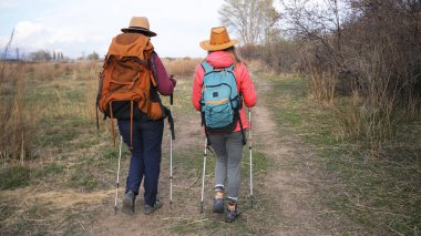 Sırt çantalarıyla toprak bir pistte yürüyen iki yolcunun arka görüntüsü. Uzun zamandır beklenen bir tatil. Aktif spor ve sağlıklı bir yaşam tarzı. Seyahat, yürüyüş ve özgürlük.