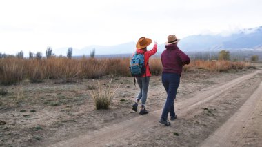 İki kadın toprak bir yolda yürüyor, biri gökyüzünü işaret ediyor. Sahne huzurlu ve huzurlu, gezginler doğa yürüyüşünün tadını çıkarıyorlar. Dağlara bir yolculuk.