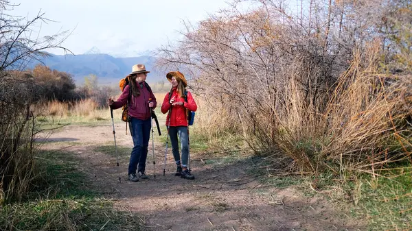 İki turist toprak bir yolda duruyor ve birbirleriyle sohbet ediyorlar. Sırt çantaları ve yürüyüş direkleri olan iki kadın dağlık yabanda uzun bir yürüyüşten sonra dinleniyorlar..