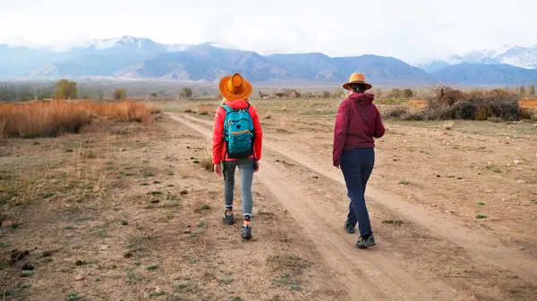 İki gezgin dağlara doğru yabanda geziniyor. Çölde toprak yolda yürüyen iki kadın, arkadan manzaralı. Gezinti, yürüyüş, seyahat