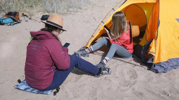 İki lezbiyen kız arkadaş sahilde çadırın yanında oturmuş telefonlarıyla sohbet ediyorlar. Açık hava tatilleri. Turistler sahilde dinlenir. Seyahat ve özgürlük