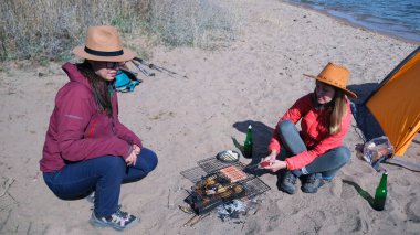 İki kız arkadaş sahilde oturuyor ve kamp ateşinin üstünde ızgarada sosis kızartıyorlar. Deniz kıyısında piknik. Çadırlı turistler hafta sonlarını aktif olarak geçiriyorlar.