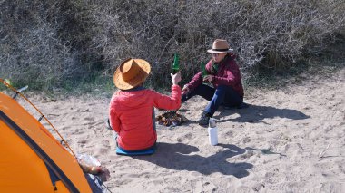 Kumsalda oturmuş bira içen iki kadın. Kamp ateşinde sosis kızartıyorlar ve sahilde çadırın yanında eğleniyorlar. Aktif hafta sonları. Özgürlük ve macera